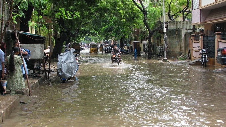 A motorbike rides through a flooded road. A woman tries to cross the road from the left hand side pavement.