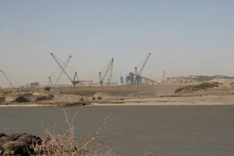 Cranes tower over the construction of a dam, with a body of water in the foreground
