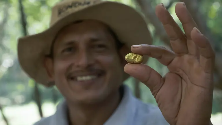 Man smiling broadly holds up to the camera a fragment of gold between his thumb and finger