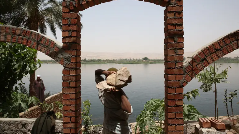 A man, his head hidden by the two large rocks he carries on his shoulders, on the edge of a large river 