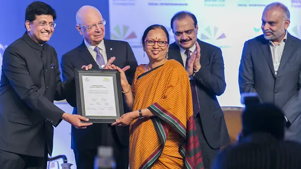 Prema Gopalan stands centre stage in an orange sari. Two men present her with a certificate; two others stand behind and applaud.
