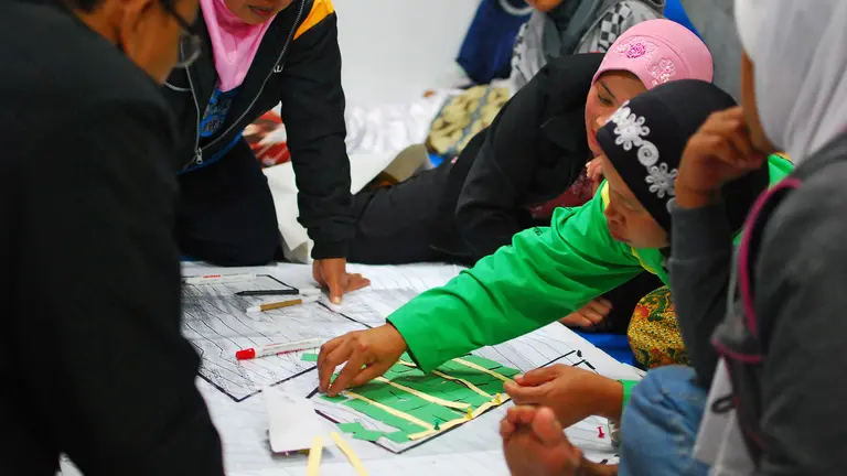 A group of men and women lean over a table and discuss the maps placed there.