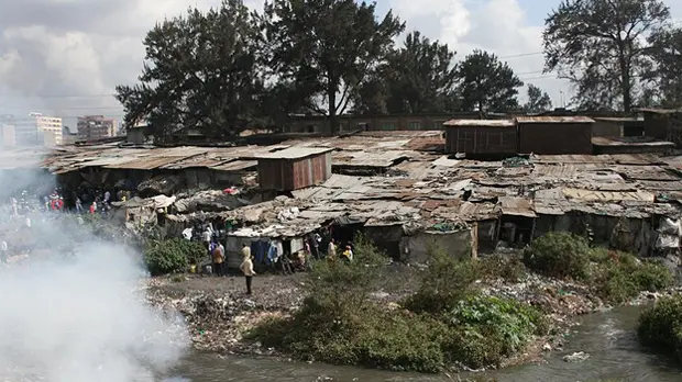 Closely built shacks. In the background, trees. In the foreground, a river and, on the other side of it, smoke.