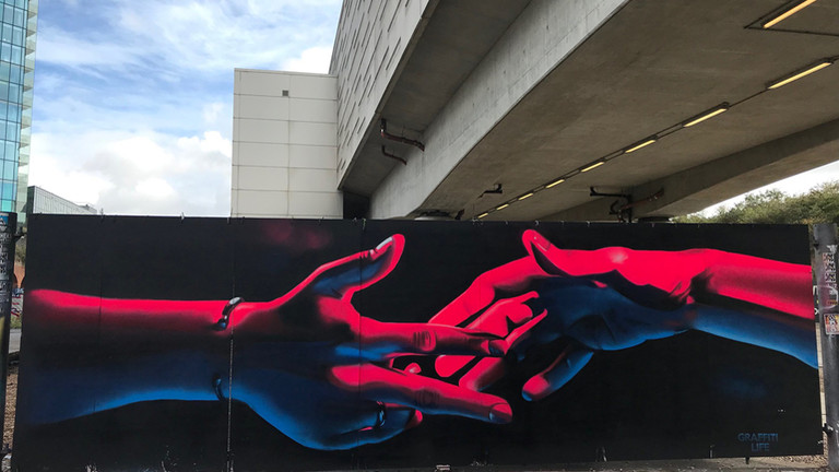 A bleak urban landscape. In the foreground, a painting of two hands reaching towards each other, painted in red and blue on a black background.