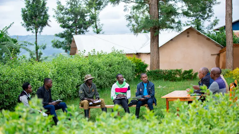 Five young men sit on benches on a lawn outside a building. They are listening to two older men seated at a desk.