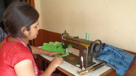 A young woman sits working at a sewing machine in a corner of a room.