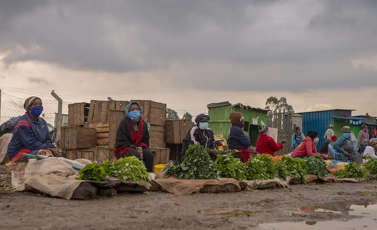 Women sitting next to vegetables wear masks and keep social distancing.