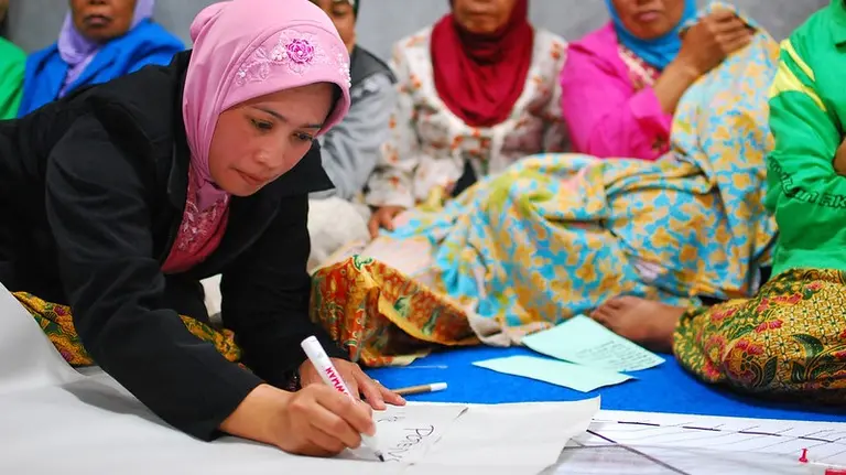 A woman writes on a poster. More women appear blurred in the background.
