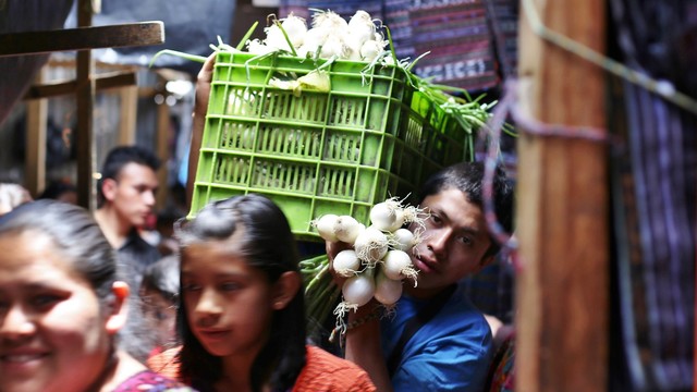 A man carries a box with spring onions through a busy scene