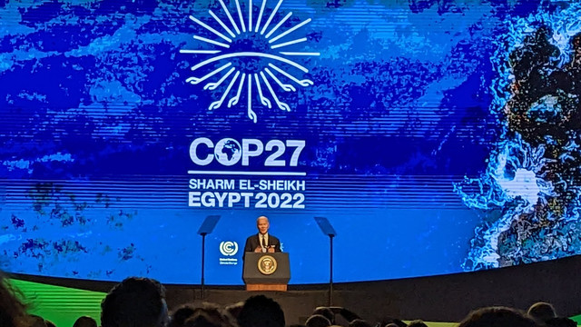 A man in the distance stands behin a podium giving a speech. Behind him, in large letters, is the COP27 logo.
