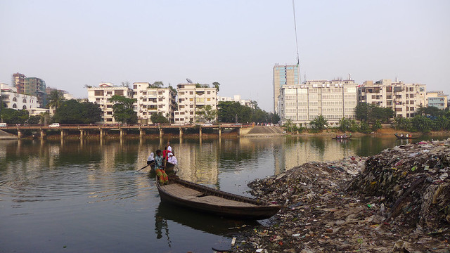 A boat on a river next to a pile of garbage with buildings in the background.