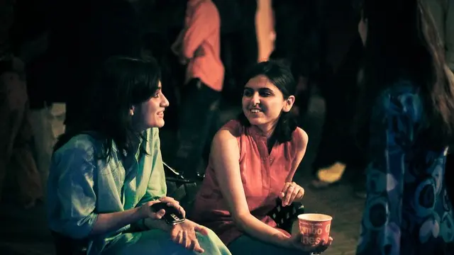 Two women chat to each sitting on a bench at night.
