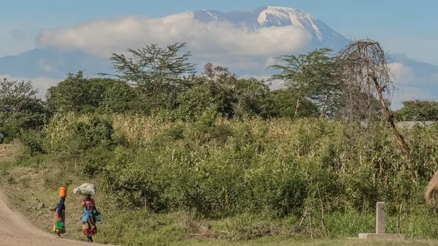 Two women walk along a road beside green undergrowth and fields, with a mountain in the background entering the clouds.