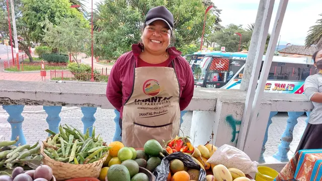 A woman stands behind a market stall of fresh vegetables, smiling proudly.