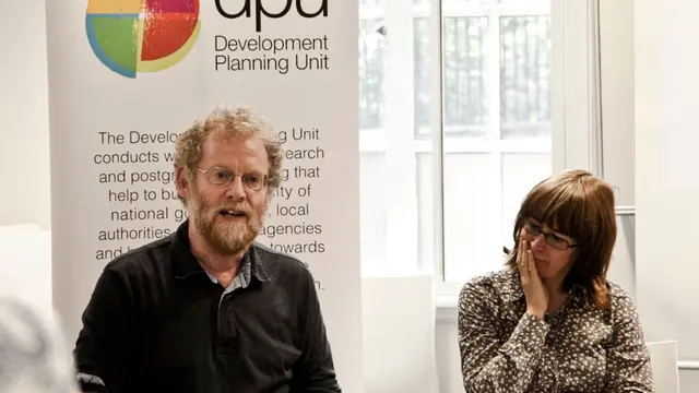 A seated man talks to a group, while a women alongside watches on. Behind them is a board advertising DPU - the Development Planning Unit at UCL.
