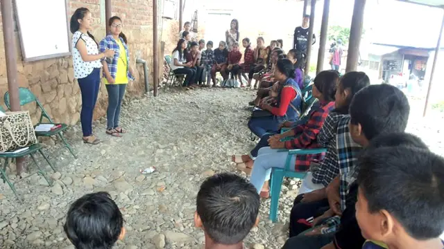 Two people stand in an outside area, surrounded by seated youngsters in a semi-circle.
