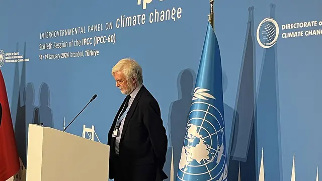 A man stands, head slightly bowed, at a podium to give a speech, with a blue IPCC flag behind him.