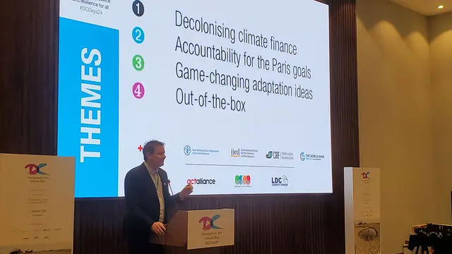 A man makes a speech at lectern in front of a big screen and surrounded by branding for Development & Climate Days.