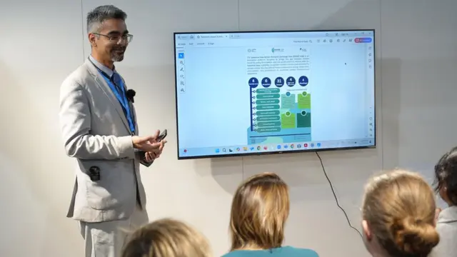 Man wearing light coloured suit presenting in front of a screen to a room of people sitting down.