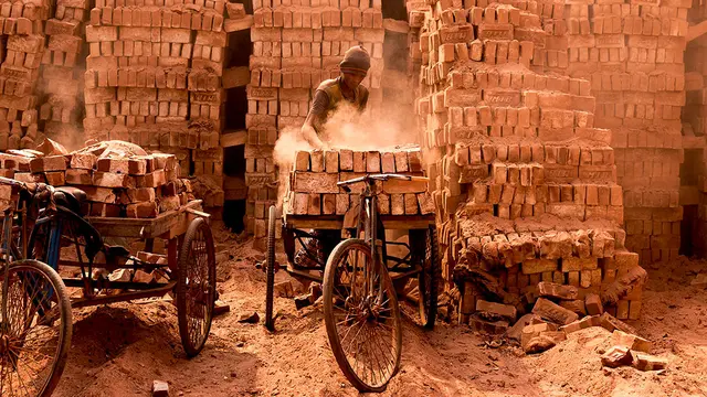 A man forges bricks in a kiln on the back of a bike, with mountains of bricks stacked up behind him.