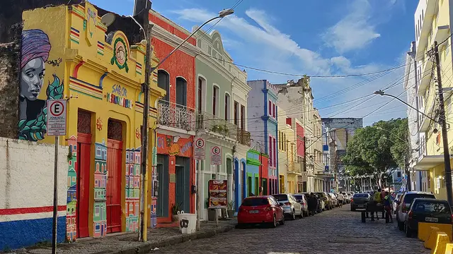 Colourful murals adorn similarly bright-coloured buildings alongside a street, under a blue sky,