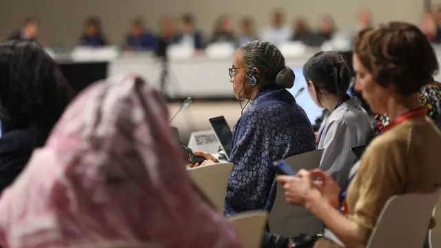 A number of seated women, one sitting behind a country sign, another holding a mobile phone, take part in a negotiation discussion.