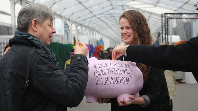 A woman holds a pink piggy bank in front of her, with the words 'Deliver climate finance' written on the front.