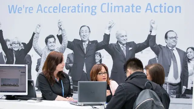 Two people taking to representatives at the Paris Agreement stand. Behind them on the wall, there is a big image of people holding hands with the words 'We're accelerating climate action'.