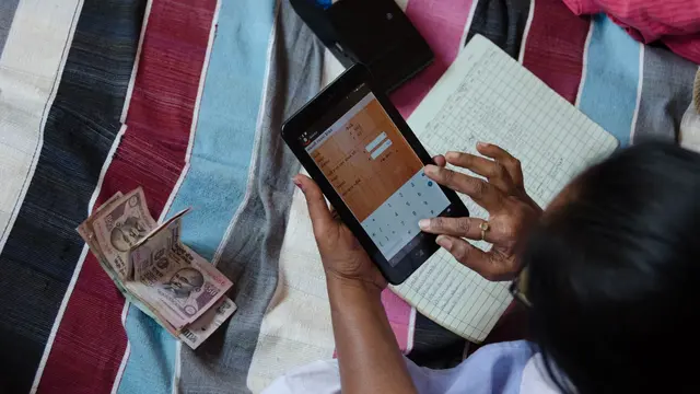 Woman holding tablet open to the calculator app, with money and a notebook besides her.