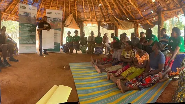 A group of seated people gather around a man who stands while he makes a presentation. They are in a hut.  