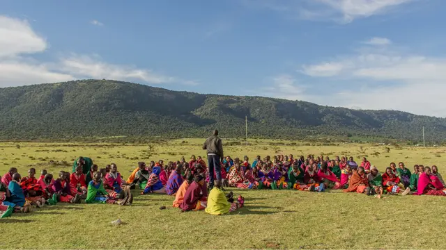 Group of women and young girls dressed in traditional clothing, sitting down in a wide open field, in a circle. Another person is standing and talking to them.