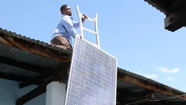 A man on top of a rook hauls up a solar panel.