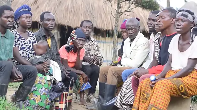 A group of Indigenous Pygmy men and women sit in a semi-circle with frustrated expressions. One woman carries a baby.