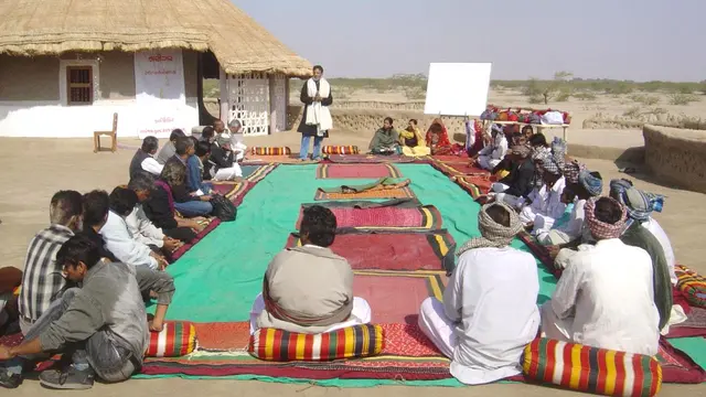 Group of community members gathered and sitting on the group, while one person is standing at the front, talking.