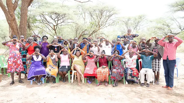 A group of people, standing and seated outside under a tree, make the heart shape with their hands as they pose for a group photo.