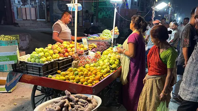A fruit vendor stands behind a wooden cart at a brightly lit night market, with shoppers gathered around