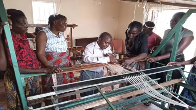 People gather around someone using a weaving machine as he demonstrates its use.