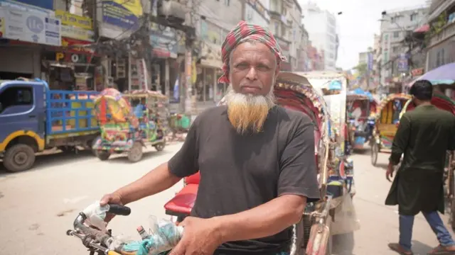 Older man standing in a middle of a busy street in Dhaka, looking straight at the camera. He is wearing a grey t-shirt and a red scarf on his head, and holding his motorcycle.