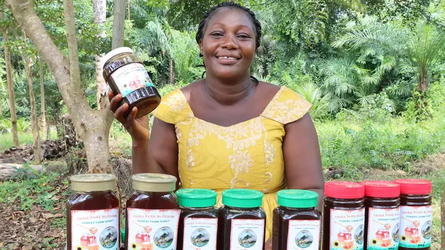 A woman sits behind stacked pots of honey, and smiles as she holds one pot in her hand.