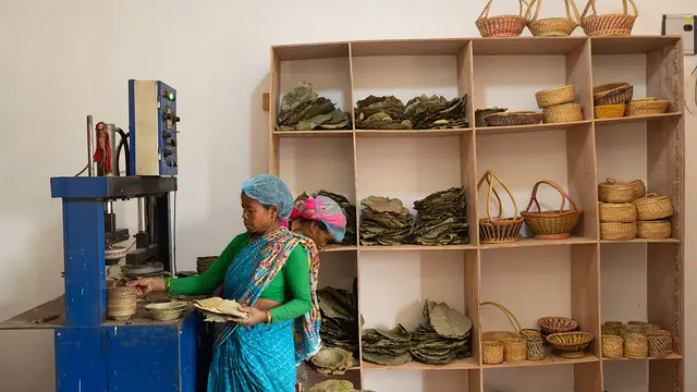 A woman stands at a machine to handcraft a basket, in front of shelves displaying lots of different types of handmade baskets.