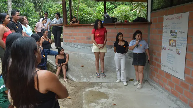 Three women make a presentation in front of a group of people, standing by a wall on which a large white sheet of paper has been stuck. 
