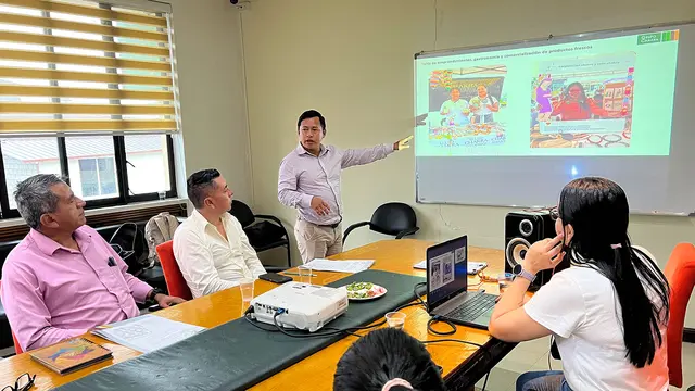 A man in an office stands in front of a screen on which a computer screen is projected and presents to a group of people around a table.