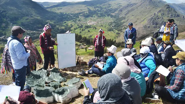 A group of people on the edge of a mountain gather around a man making a presentation and holding a large sheet of white paper, with a spectacular mountain backdrop.