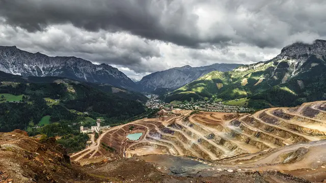 A long-range photo of a landscape showing tiered levels that denote mining in front of mountains.   