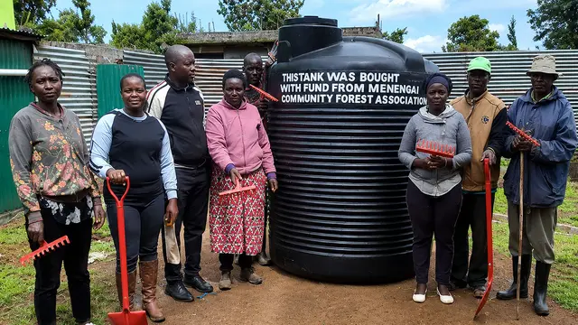 A group of men and women pose for a photo either side of a person-high large black water tank.