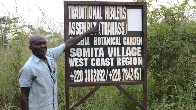 A man standing outside with bushes behind points to a large black sign on which there is white writing stating: "Traditional Healers Assembly"