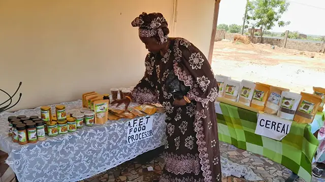 A woman bends over a table stocked with small pots/products to sell. A table behind is labelled 'cereal'.
