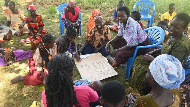 A group of people, sitting in chairs and on the ground in a circle, write on a piece of paper on the ground in the middle. 