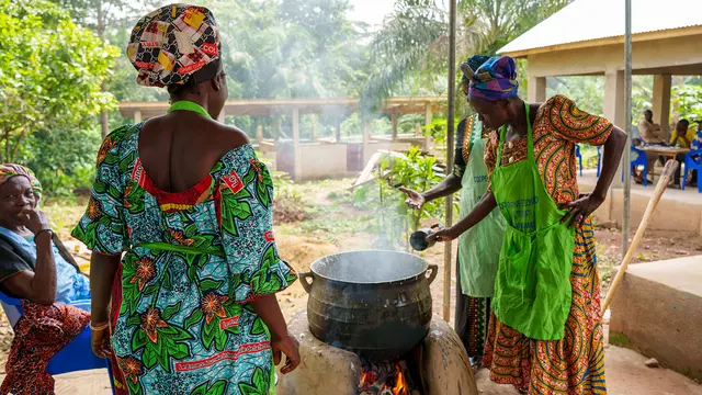 Women in brightly coloured clothes gather around a large smoking pot outside.