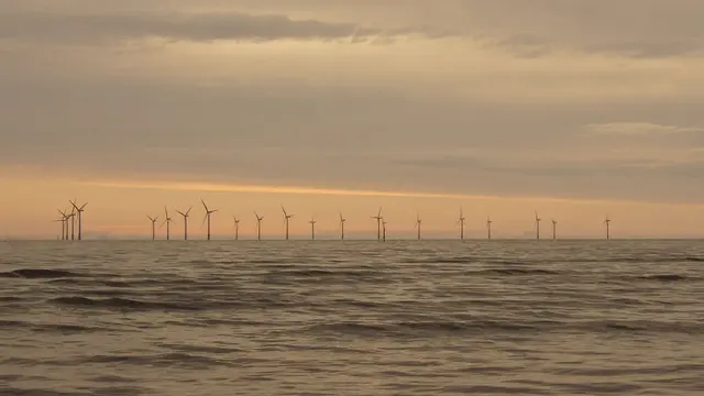 A dramatic sunset of reds and yellows frames a dozen wind turbines in the distance in the water.